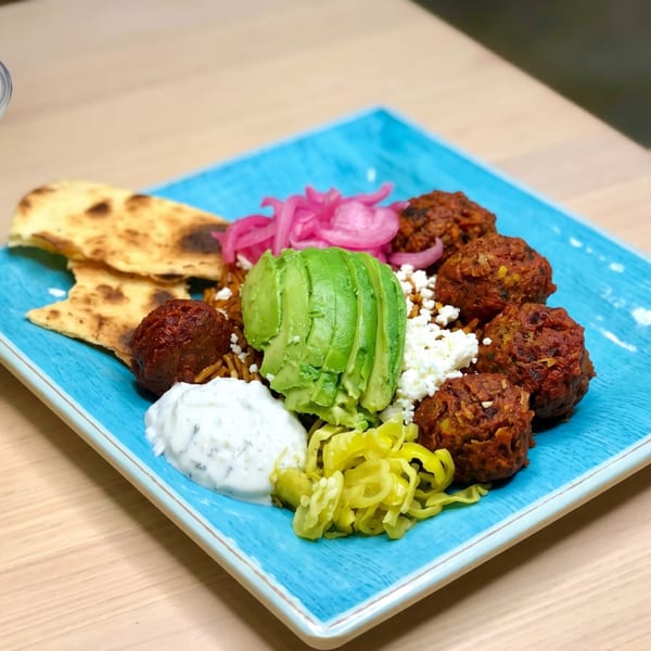 A vibrant blue plate holds falafel, pita bread, avocado, pickled onions, feta, and tzatziki sauce.