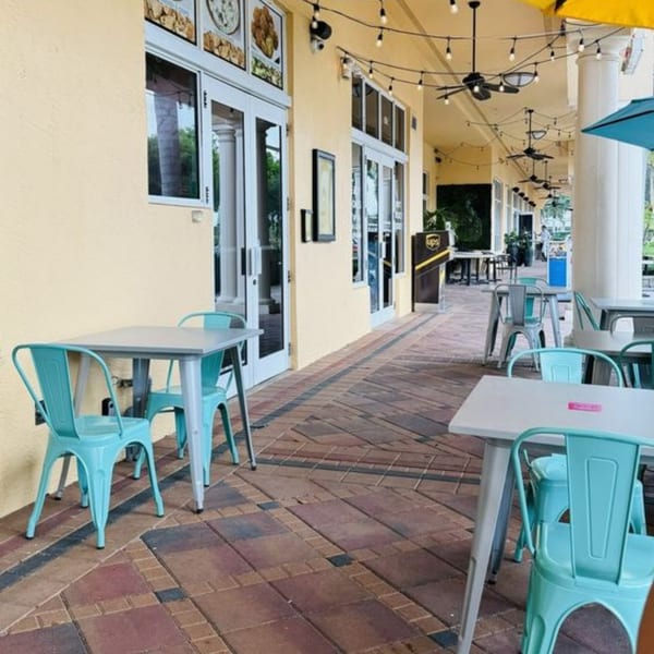 Outdoor patio with light blue metal chairs and gray tables along a yellow building with string lights.