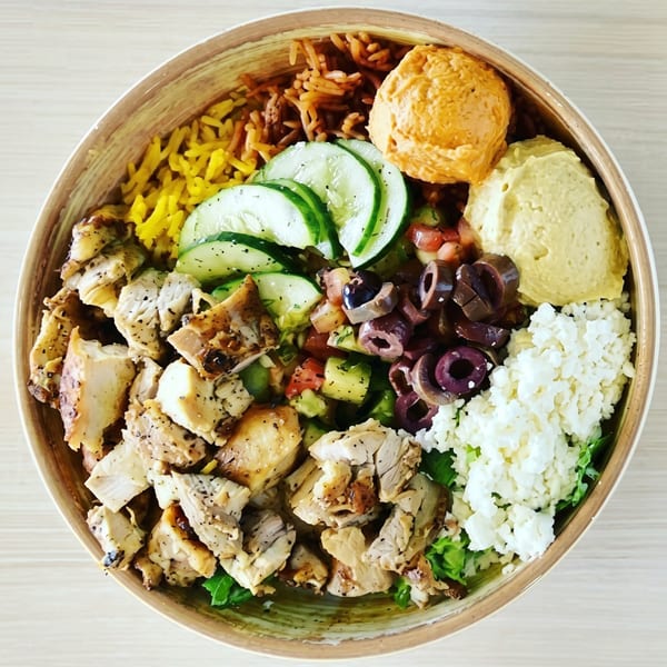 A colorful bowl of Mediterranean food with chicken, rice, hummus, cucumbers, olives, and feta.