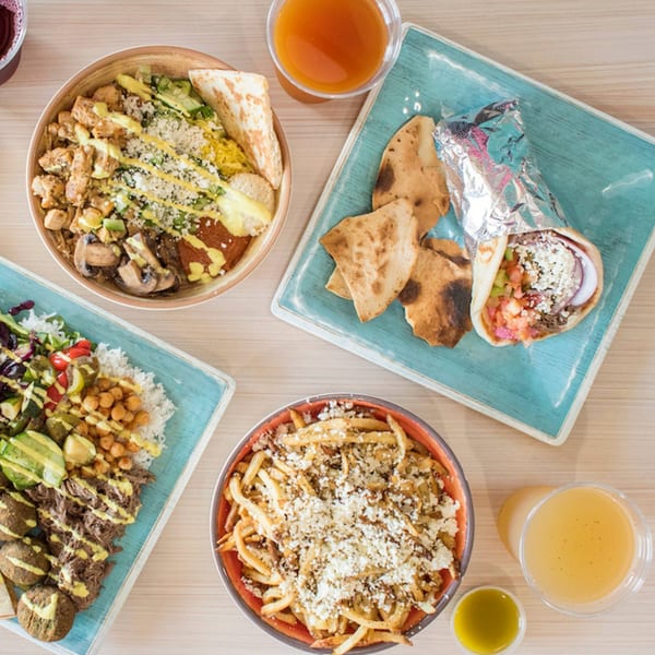 Overhead view of various Mediterranean dishes, including bowls, a wrap, and fries, on a light wooden table.