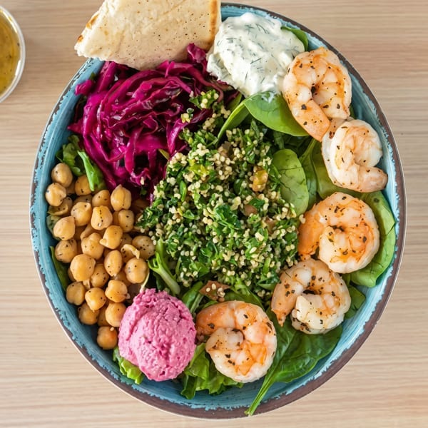 Colorful Mediterranean bowl with shrimp, chickpeas, tabbouleh, red cabbage, hummus, and pita bread.