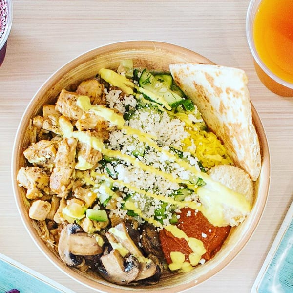 A colorful bowl with chicken, mushrooms, rice, vegetables, sauces, and pita bread.
