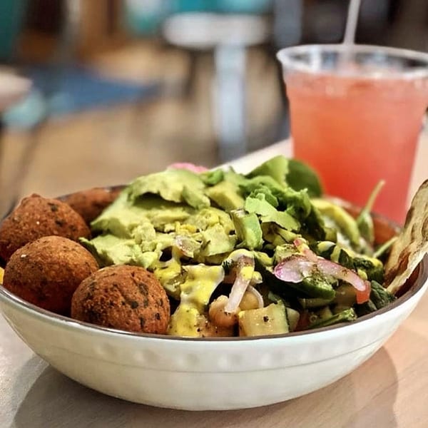 A white bowl filled with falafel, avocado, salad, and a pink drink in the background.