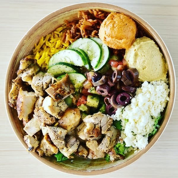 Overhead view of a colorful Mediterranean bowl with chicken, rice, hummus, cucumbers, olives, and feta.