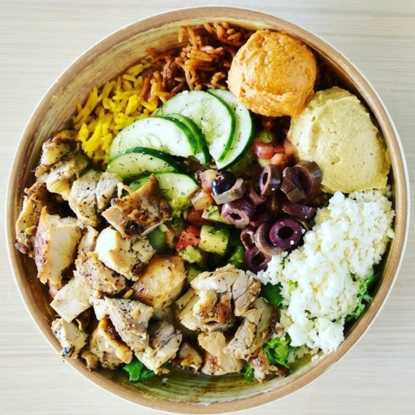 Overhead view of a colorful Mediterranean bowl with chicken, rice, hummus, cucumbers, olives, and feta.