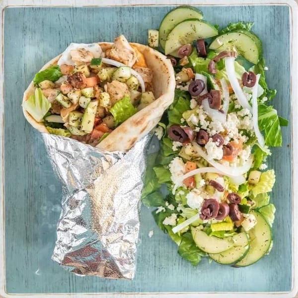 A chicken pita wrap in foil next to a Greek salad with olives, feta, and cucumbers.