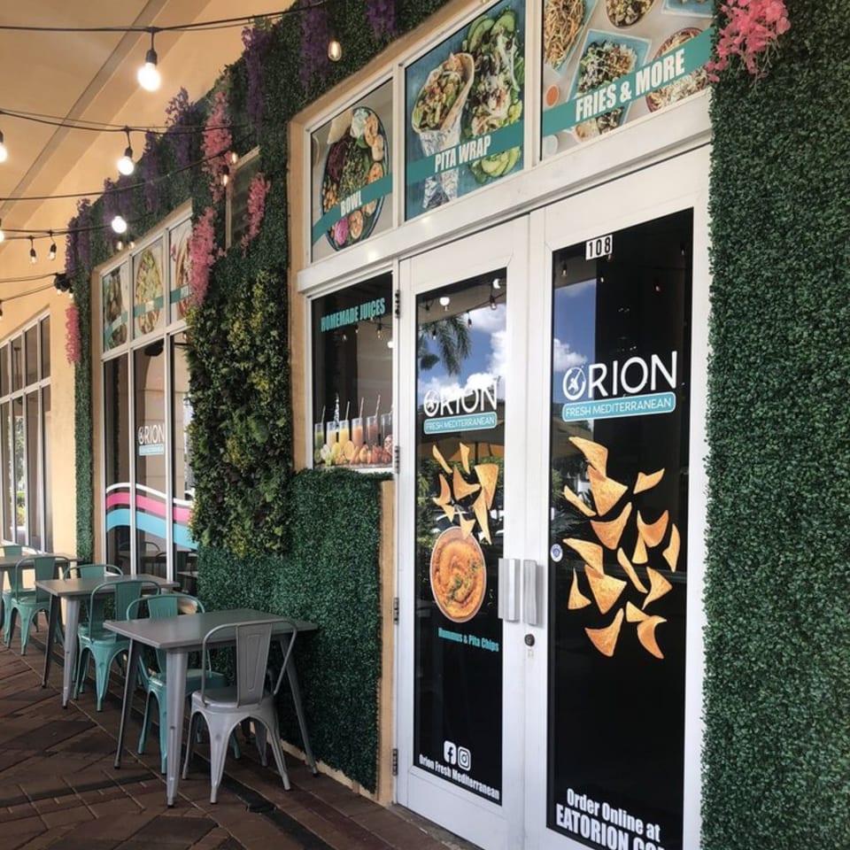 Orion Fresh Mediterranean restaurant exterior with outdoor seating, green foliage walls, and menu displays.