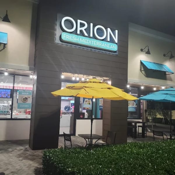 Exterior of Orion Fresh Mediterranean restaurant at night with outdoor seating under yellow and blue umbrellas.