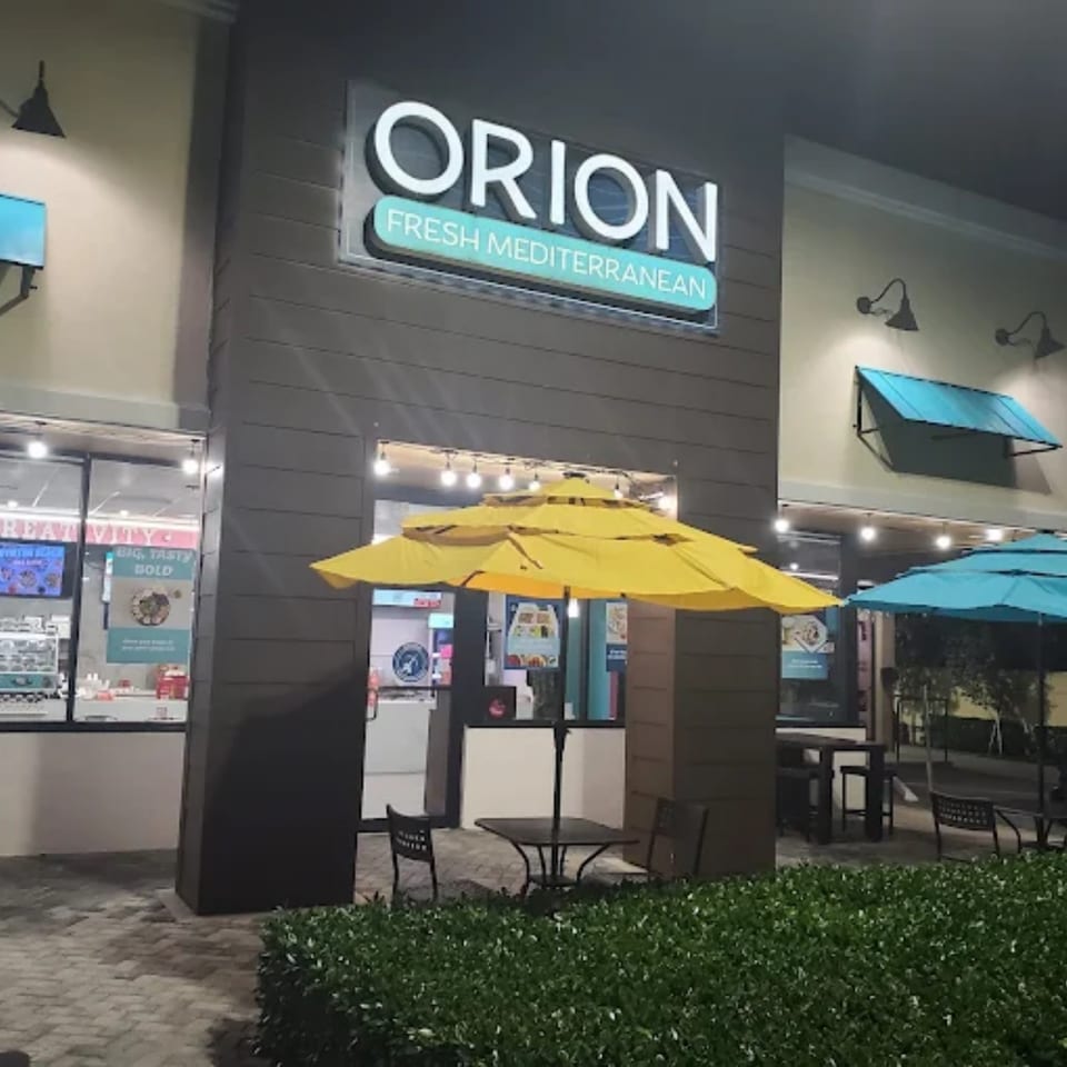 Exterior of Orion Fresh Mediterranean restaurant at night with outdoor seating under yellow and blue umbrellas.